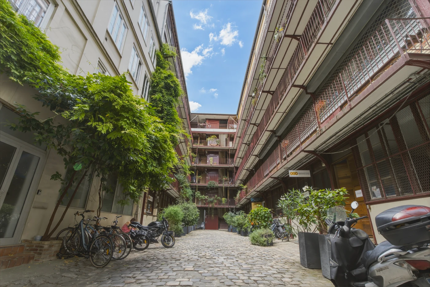 Cour pavée et végétalisée entre des façades à coursives métalliques, sous un ciel bleu
