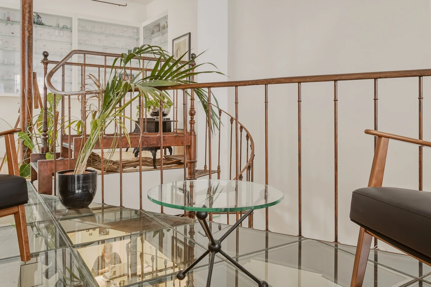 Escalier à rampe métallique, sol en verre, table ronde et plante dans un intérieur lumineux
