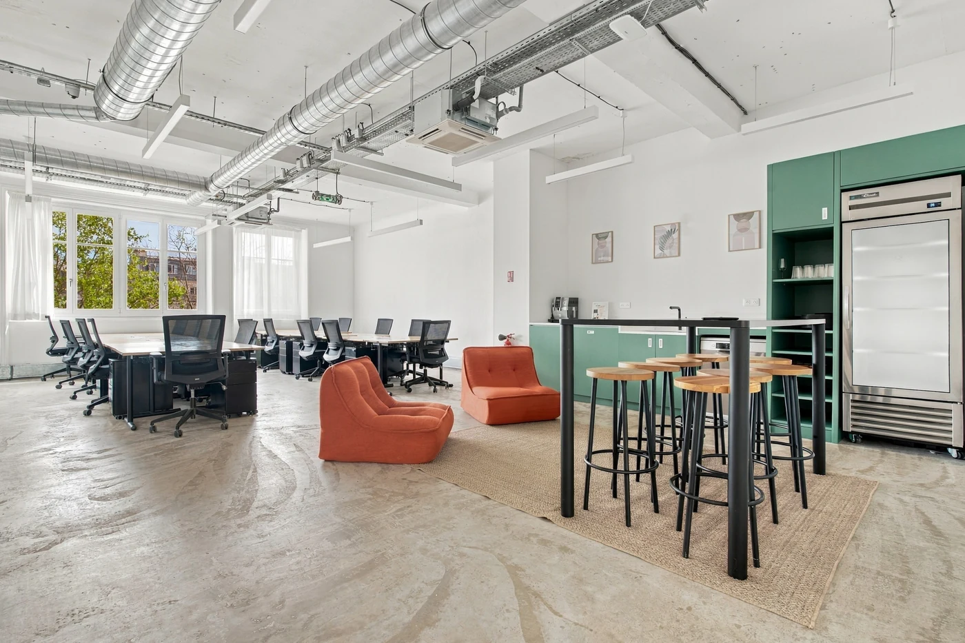 Espace de travail partagé avec postes de bureau, fauteuils orange et cuisine verte sous un plafond industriel