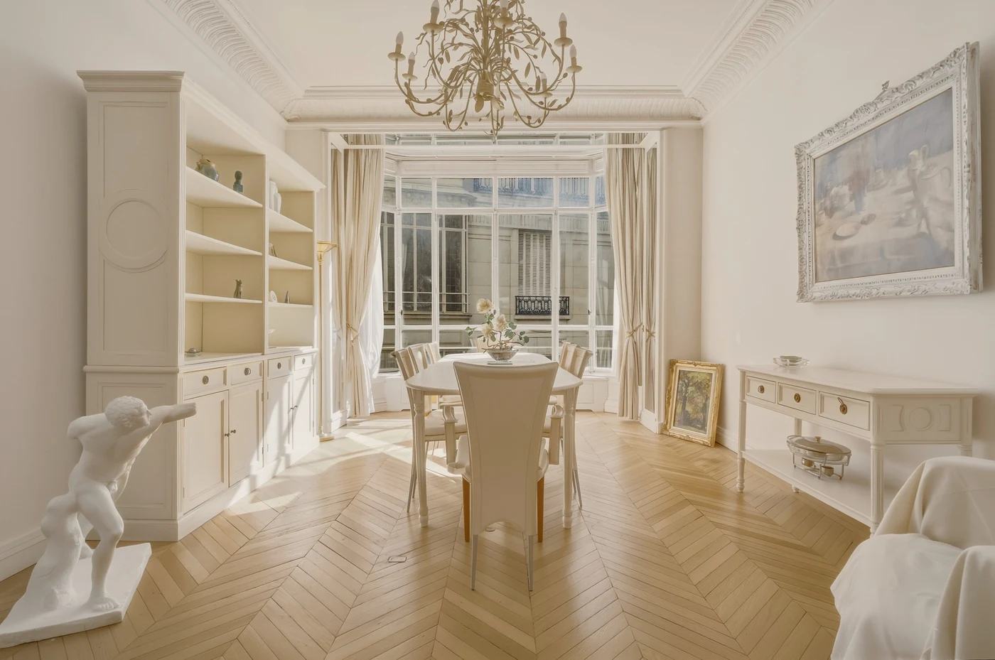 Salle à manger aux tons crème, lustre, bibliothèque et parquet éclairé par les fenêtres